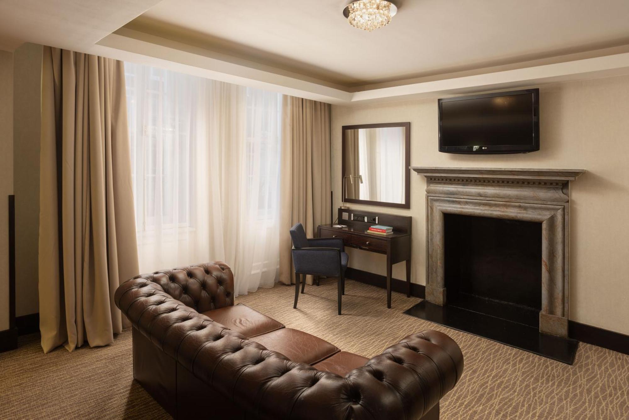 Hotel Montcalm Brewery Townhouse London Exterior foto The photo shows a stylish interior of a room, likely a living area or a hotel suite. It features a dark leather chesterfield sofa with tufted upholstery. Opposite the sofa is a small wooden desk with a chair, positioned near a wall-mounted flat-scree