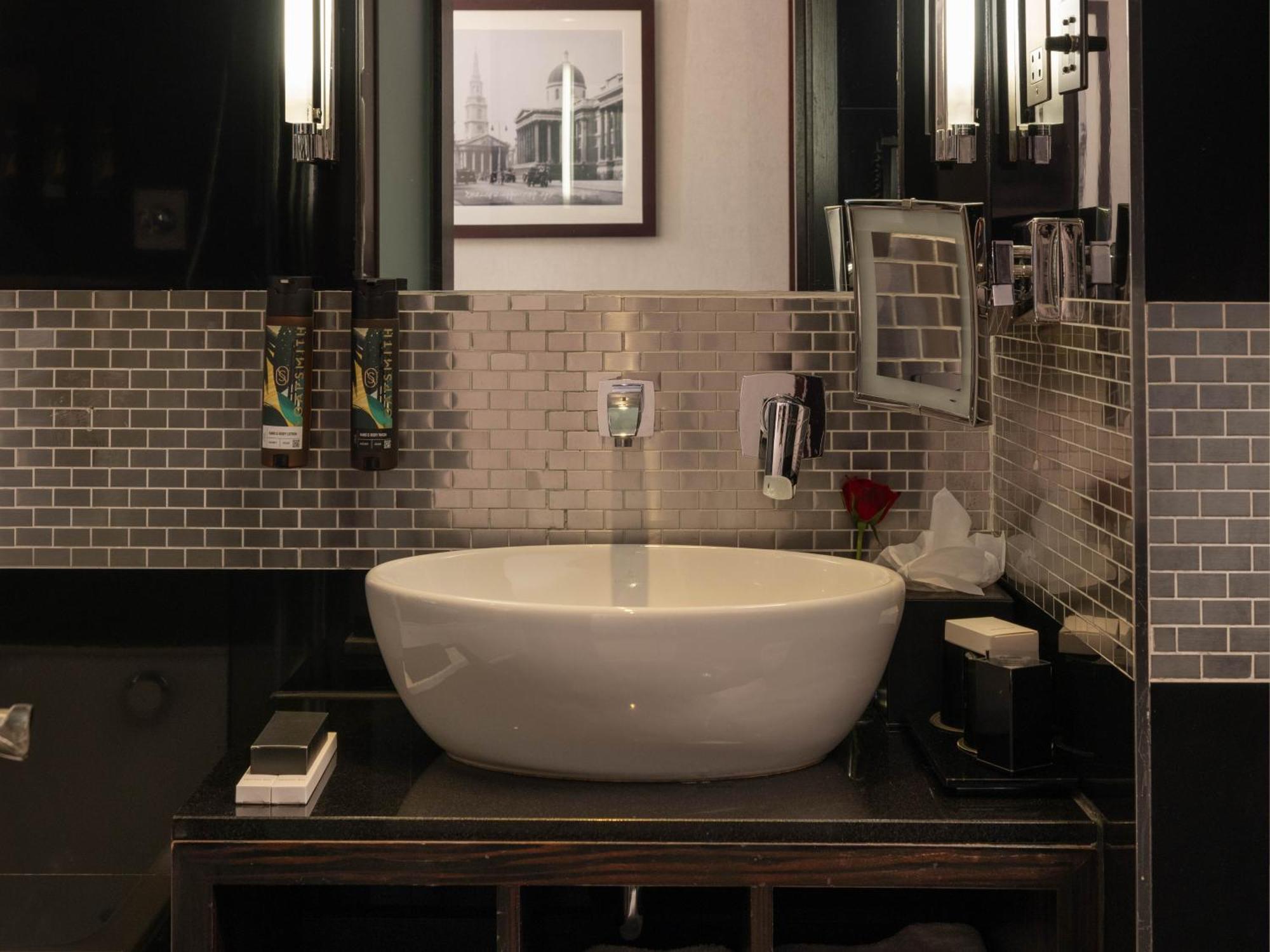 Hotel Montcalm Brewery Townhouse London Exterior foto The photo shows a modern bathroom with a stylish design. It features a large, round white sink placed on a dark wooden vanity. The walls are adorned with a mix of gray and silver tile, creating a sleek and contemporary look. Above the sink, there is 