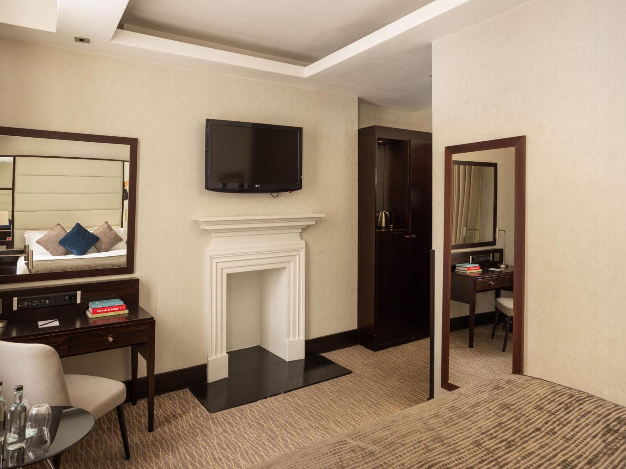 Hotel Montcalm Brewery Townhouse London Exterior foto The photo depicts a stylish hotel room. It features a double bed, a large flat-screen TV mounted on the wall, and a decorative fireplace. There is a wooden desk with a chair beside it, as well as a mirror hanging on the wall. The decor is modern and 