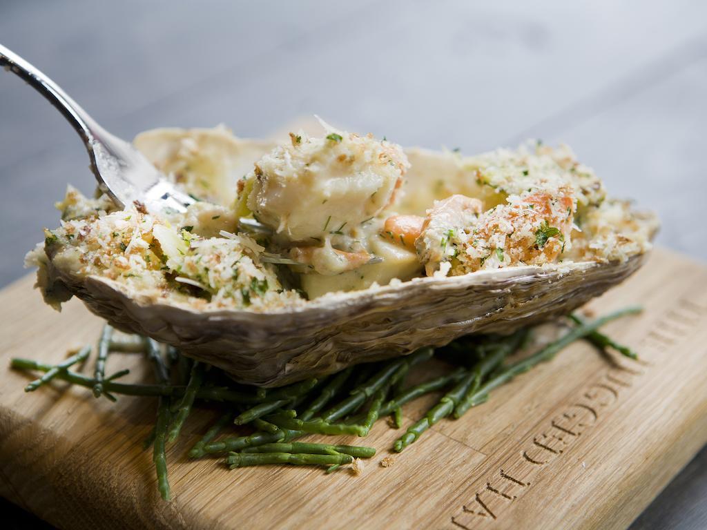 Hotel Montcalm Brewery Townhouse London Exterior foto The photo shows a large oyster shell filled with a mixture that appears to include seafood, herbs, and breadcrumbs. The dish is presented on a wooden board with some greenery, possibly a type of seaweed or garnishing. A fork is placed beside the oyst