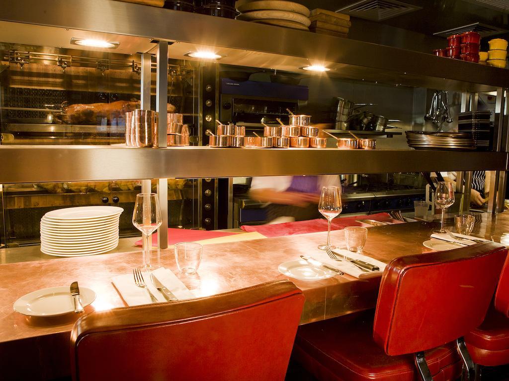 Hotel Montcalm Brewery Townhouse London Exterior foto The photo shows a restaurant interior featuring a bar or counter area. There are red chairs arranged around a copper-topped table where dining utensils and plates are set. Above the counter, there are shelves displaying various cooking pots and kitch