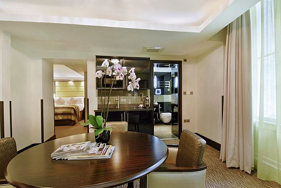 Hotel Montcalm Brewery Townhouse London Exterior foto The photo shows a stylish hotel room or suite. In the foreground, there is a round dining table with a few magazines on it. Surrounding the table are upholstered chairs. To the right, there are large windows with light-colored curtains, allowing natu