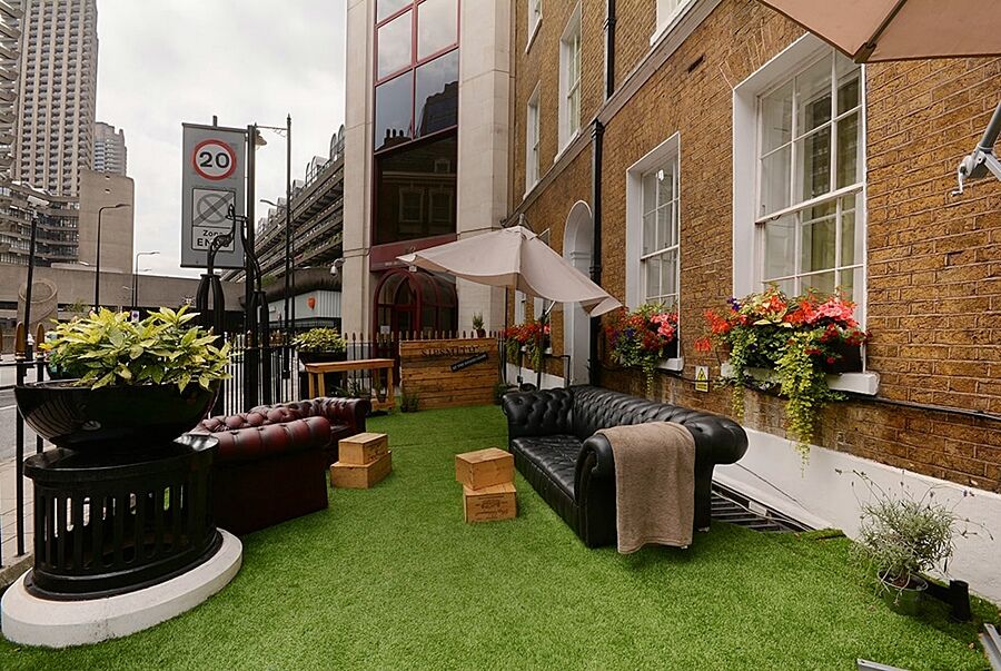 Hotel Montcalm Brewery Townhouse London Exterior foto The photo depicts a charming outdoor space, likely a patio or garden area. It features two comfortable-looking leather sofas, one of which is a deep burgundy color, placed on a green artificial lawn. There are wooden crates that could serve as side t